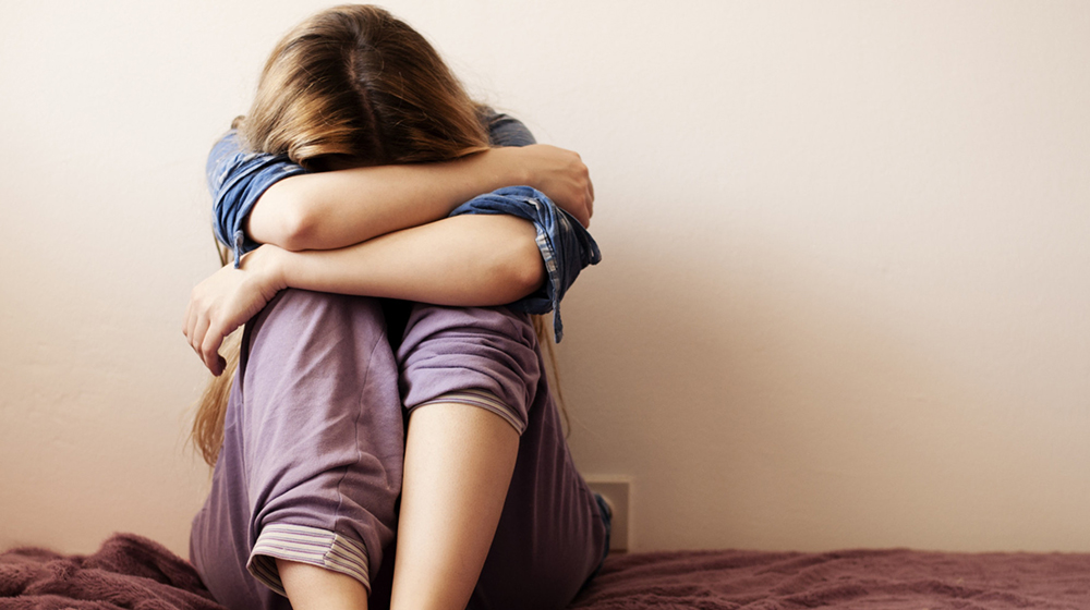 Person sitting on a bed with arms wrapped around their knees and head down, conveying distress or sadness
